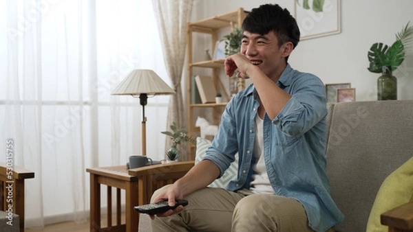 Fototapeta leisure Korean young man with tv controller is laughing and slapping on his lap while watching a funny comedy show in the living room at home during daytime