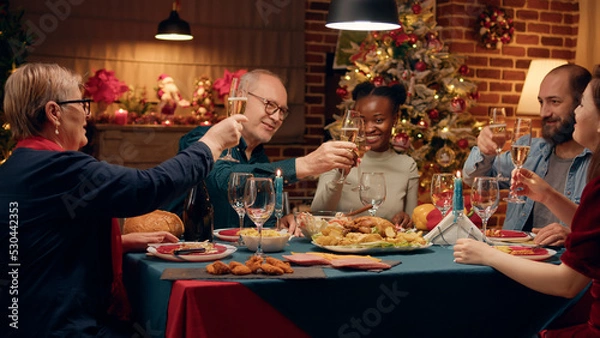 Obraz Happy senior man inviting family members to toast while celebrating winter holiday at home. Festive relatives gathered to celebrate Christmas with traditional home cooked food and sparkling wine.