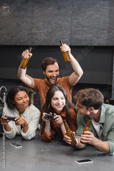 Fototapeta KYIV, UKRAINE - JULY 26, 2022: happy interracial women holding joysticks near friends with beer in kitchen