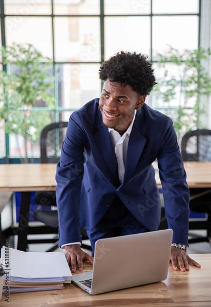 Fototapeta African-American office worker standing and leaning on desk. Vertical shot of concentrated young male employee working on laptop in modern office. Occupation, emotion, business concept