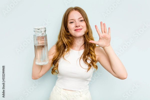 Fototapeta Young caucasian woman holding jar of water isolated on blue background smiling cheerful showing number five with fingers.