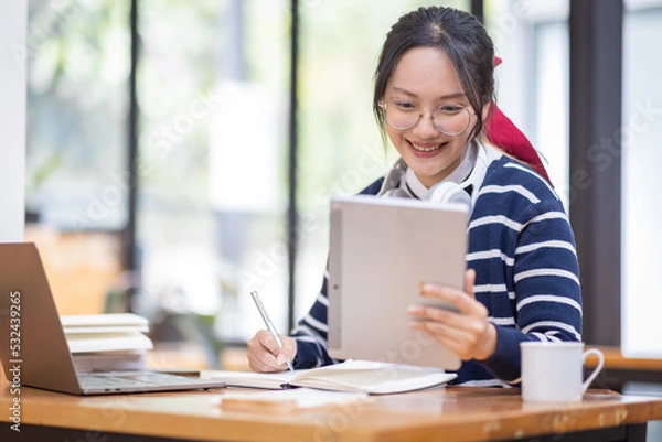 Fototapeta Young adult happy smiling Hispanic Asian student wearing headphones talking on online chat meeting using laptop in university campus or at virtual office. College female student learning remotely.
