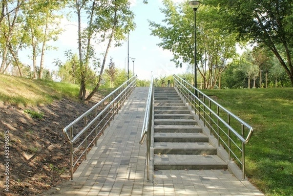 Fototapeta Stairs and ramp with metal railings for the passage of strollers and wheelchairs in public park. Concept of comfortable barrier-free urban environment in city. Pathway for people with disabilities