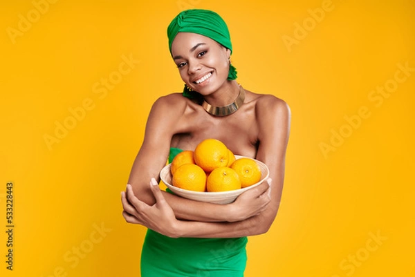 Fototapeta Attractive African woman in traditional headwear holding oranges and smiling against yellow background