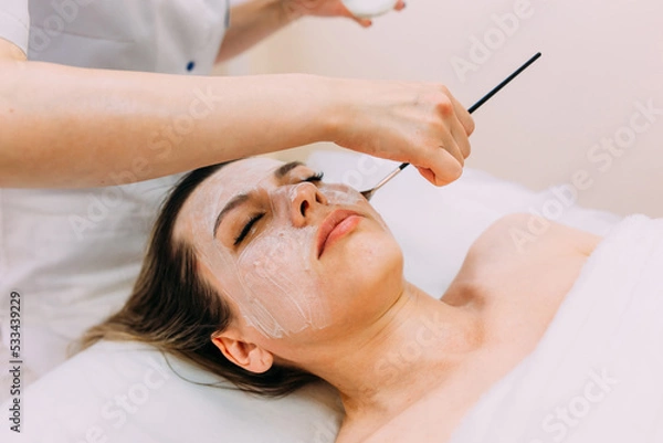 Fototapeta Cosmetologist makes cleansing peeling and face mask for woman. Beauty skin care