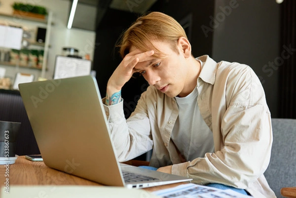 Fototapeta Portrait of tired upset exhausted man sitting in front of laptop with hand over forehead, having headache, trying to concentrate and read information on screen. Freelance and deadline