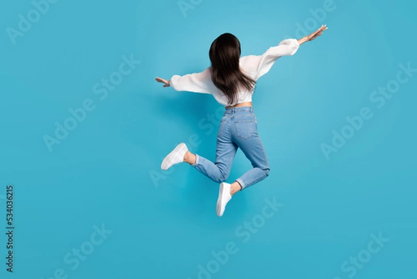 Fototapeta Full size rear behind photo of overjoyed active girl arms wings flying jump isolated on blue color background