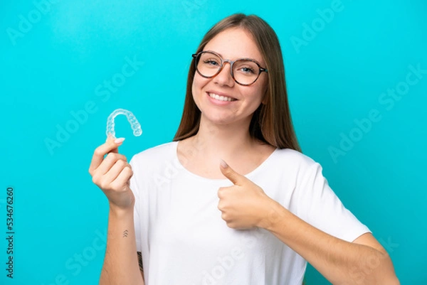 Fototapeta Young Lithuanian woman holding invisible braces isolated on blue background with thumbs up because something good has happened