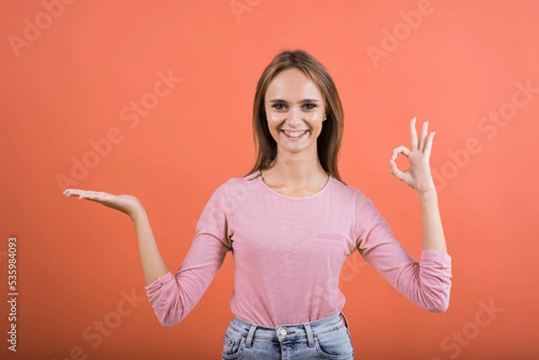 Fototapeta The girl shows an Okay gesture and holds on a palm a copy space for advertising on a red background in the palm of her hand.