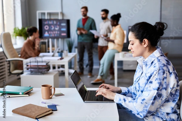 Fototapeta Side view of young serious female employee typing on laptop keypad while sitting by desk in openspace office against her colleagues