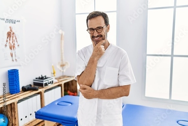 Fototapeta Middle age man with beard working at pain recovery clinic looking confident at the camera smiling with crossed arms and hand raised on chin. thinking positive.