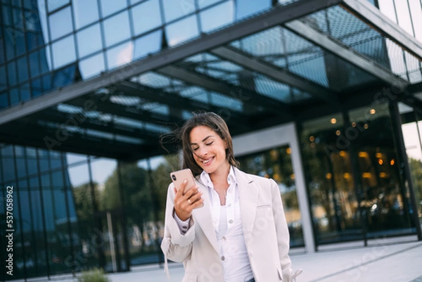 Fototapeta Young cheerful business woman working with a mobile phone in the street with office buildings in the background