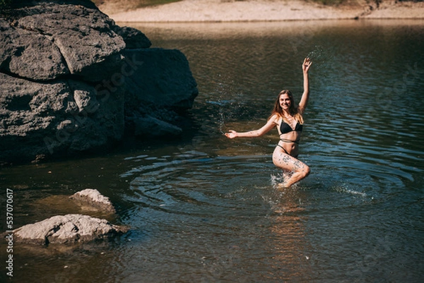 Fototapeta A young girl with long blond hair, a slender figure, in a black swimsuit jumps in the river against the backdrop of rocks and laughs.