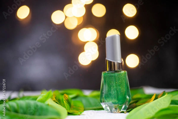 Fototapeta green nail polish on green leaves The back has a black-grey, blurry background with a soft, round orange bokeh light