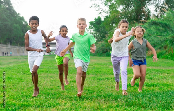 Fototapeta Glad young boys and girls running on green grass on field.