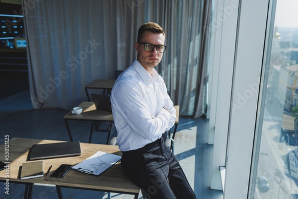 Fototapeta Cute brutal young businessman in glasses and white shirt, he is in modern office with big windows. A man in classic trousers and a white shirt. Young attractive businessman