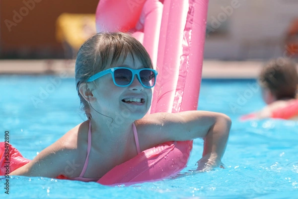 Fototapeta Young joyful child girl having fun swimming on inflatable air mattress in swimming pool with blue water on warm summer day on tropical vacations. Summertime activities concept