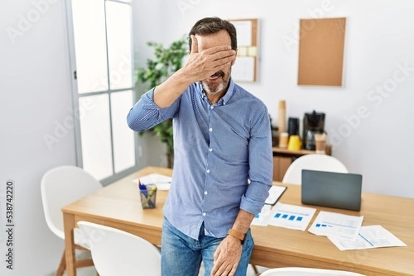 Fototapeta Middle age hispanic man with beard wearing business clothes at the office smiling and laughing with hand on face covering eyes for surprise. blind concept.