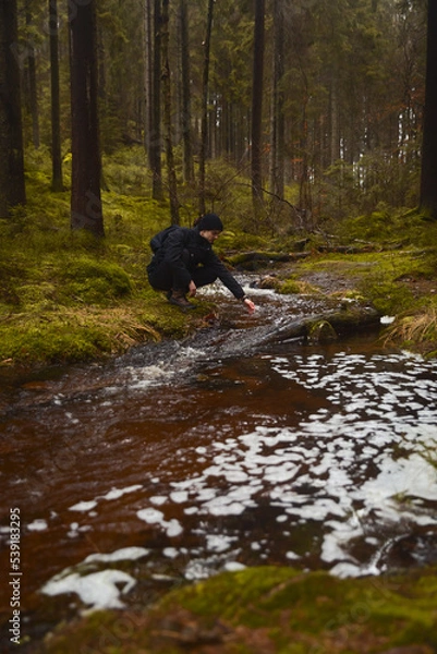 Fototapeta A caucasian man with a backpack sitting next to a stream in a forest touching the water.