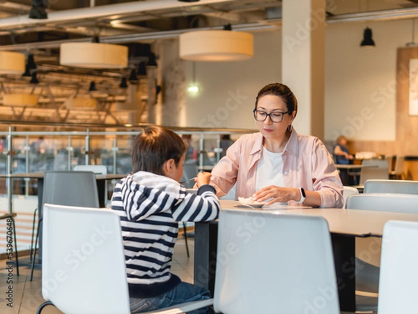 Obraz Mother and son are eating in cafe. Family on food court in shopping mall. Woman and kid have lunch in modern unrecognizable cafeteria.