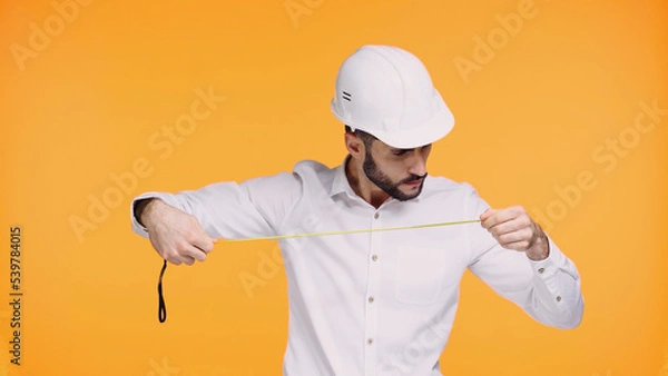 Fototapeta focused architect in hard hat looking at measuring tape isolated on yellow