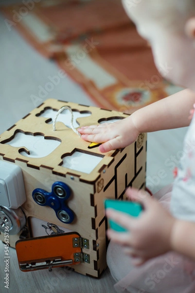 Obraz sensory board. A child is trying to find a figure for a sorter in a busy cube. A toy to improve color and shape recognition skills. Educational wooden toy for wrist coordination