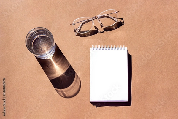 Fototapeta Empty notepad, glass of water and eyeglasses with strong shadows on brown background. Creative mockup. Top view, flat lay, Still life