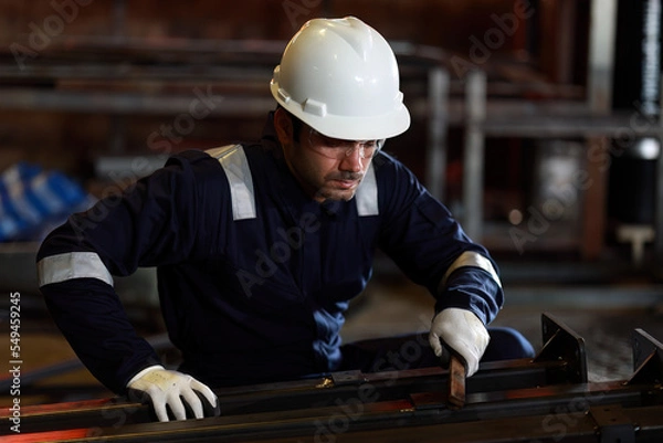 Fototapeta Skilled industrial worker finishes polishing rusty piece of metal on a table and rubs the dust with his hand. Interior of heavy industry machine building plant.