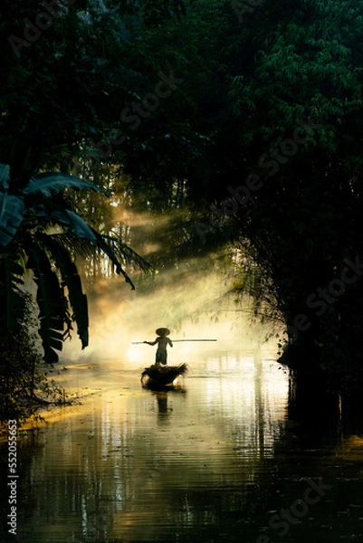 Fototapeta Photo of a local asian old man male boatman wearing conical hat rowing a small wooden boat across a small river during sunset time in a bamboo forest to deliver some dry grasses as animal feeds. 