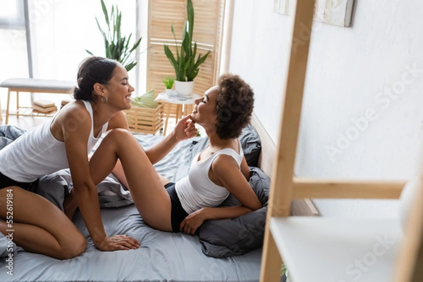 Fototapeta happy lesbian african american woman touching chin of young girlfriend in modern bedroom.