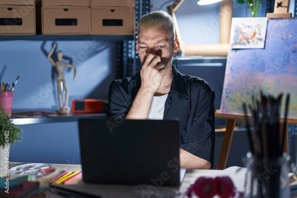 Fototapeta Young caucasian man using laptop at night at art studio smelling something stinky and disgusting, intolerable smell, holding breath with fingers on nose. bad smell