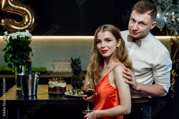 Fototapeta a married couple poses and hugs at a festive table on the occasion of a birthday;
alcohol and snacks are on the table;
glasses with red wine in hands;
boy and girl express sincere feelings