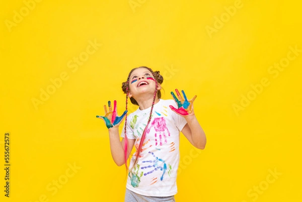 Fototapeta A little girl, painted with paints, looks at your advertisement on a yellow isolated background. The art of painting with paints for children. Art courses for schoolchildren.  Copy space.