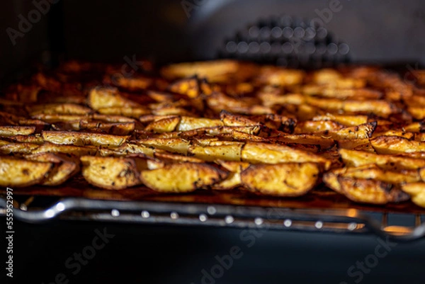 Fototapeta Crispy, golden brown baked potato wedges seasoned with paprika, garlic, salt, pepper, rosemary, basil on tray in the oven photographed from the side.