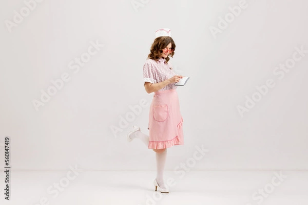 Fototapeta Emotional young woman, retro waitress in american fashion style of 70s, 80s uniform posing over light studio background. Comic photography style