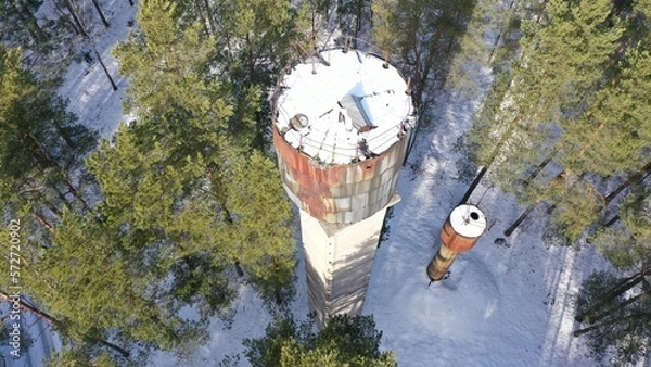 Fototapeta An old rusty water tower and a fire tower under a layer of snow in the middle of a dense spruce forest in winter. Frozen water turned into ice. Chernobyl in winter.