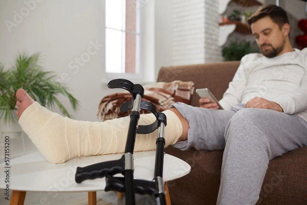 Fototapeta Young man sitting on the sofa at home with his broken injured leg in a cast or bandage resting on a foot stool, and crutches leaning on it. Injury, accident concept