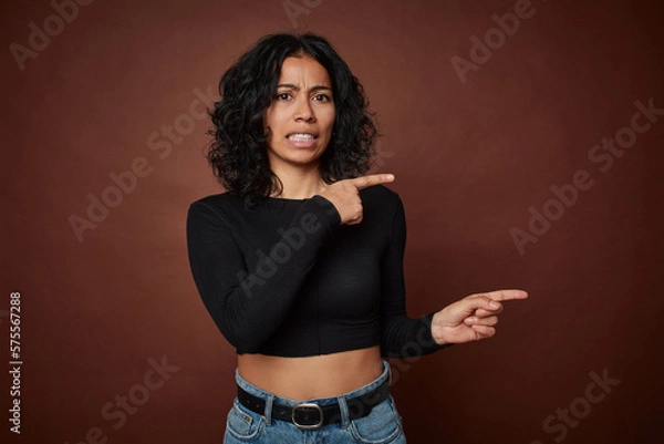 Fototapeta Young colombian curly hair woman isolated on brown background shocked pointing with index fingers to a copy space.