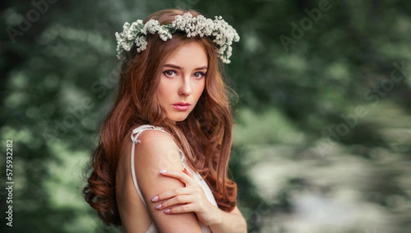 Fototapeta Beautiful red haired girl in white vintage dress and wreath of flowers standing under the tree and looking at camera. Fairytale story. Warm art work.