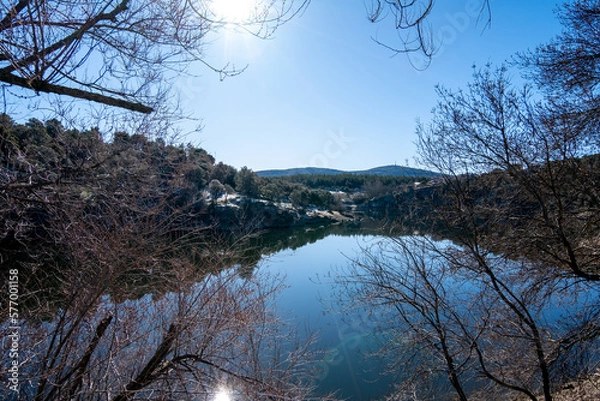 Obraz Paisaje del río en un pueblo cerca de Burgos con las montañas nevadas de fondo bajo un cielo azul soleado y las ramas de los árboles en primer plano.