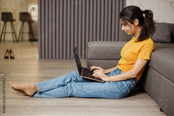 Fototapeta Happy indian or arabian young woman sitting on floor and working by laptop