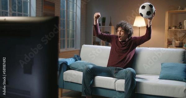 Fototapeta Expressive Spain man football team supporter emotionally watching a soccer game. Hipster guy with curly hair watching tv at home alone 