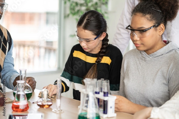 Fototapeta Diversity group of students in protective eyeglasses in science class doing chemical experiment in laboratory. Teacher watching girl mixing chemistry in test tube. learning and education concept