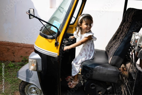 Fototapeta Caucasian girl with dark hair in a white dress poses behind the wheel of an Indian motorized rickshaw. Tourism in Asia. Children's adventures in India.Playing as a motorized rickshaw driver.Incredible