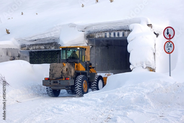 Fototapeta A large tractor clears snow from the entrance to a narrow tunnel. Snowbound narrow country road going through rectangular concrete tunnel and great snowdrifts.