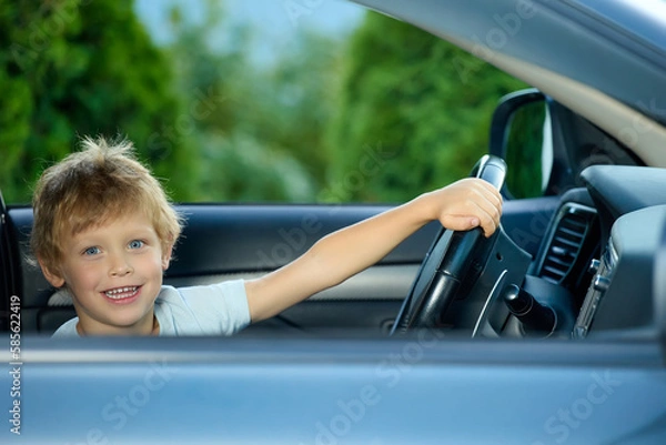 Fototapeta Cute little boy behind the wheel of an expensive car holds the steering wheel, happily looks into the camera. Fulfilling a child's dream of sitting behind the wheel in a real car