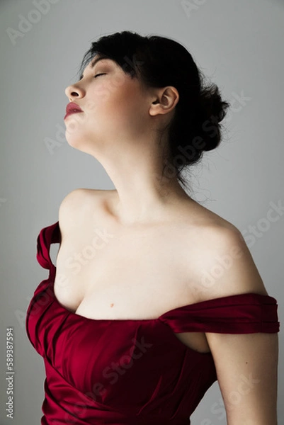 Fototapeta Portrait of a beautiful caucasian woman in her 30s wearing a red off the shoulder dress. She has dark brown hair that is in a bun. Her head is tilted up and her eyes are closed. 
Description