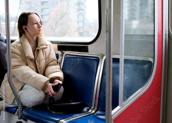 Fototapeta young girl teenager rides in transport looks out the window headphone mobile phone peaceful face space for text Sky Train bus car ordinary person in real life street Cities canada suburb surrey