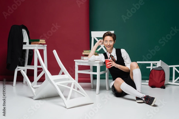 Fototapeta happy young student in eyeglasses holding drink in plastic cup near chairs and books on green and burgundy background.