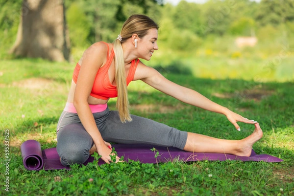 Fototapeta Image of smiling fitness woman 20s wearing headphones working out and stretching legs while sitting on exercise mat in green park